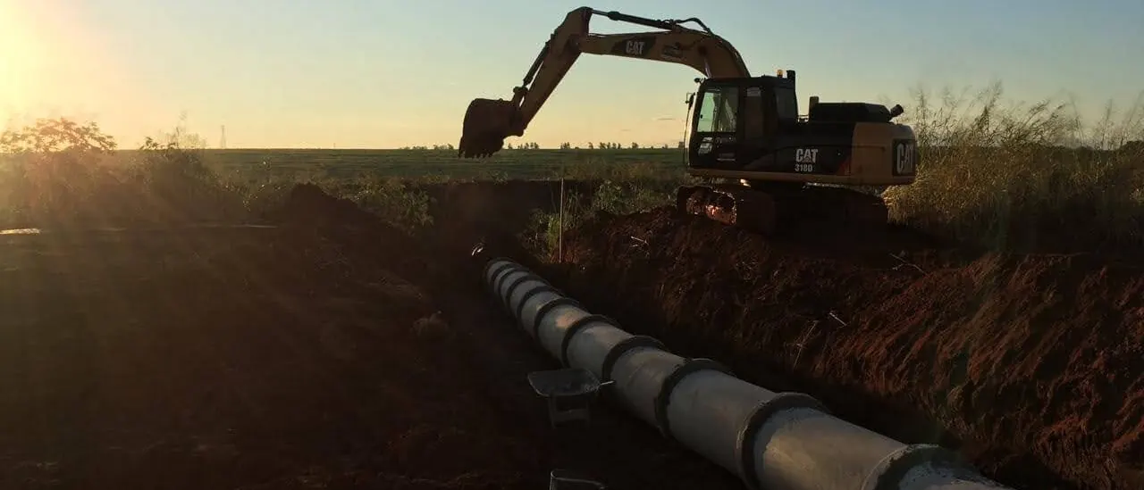Escavadeira Caterpillar ao pôr do sol sobre vala com tubos de concreto já assentados