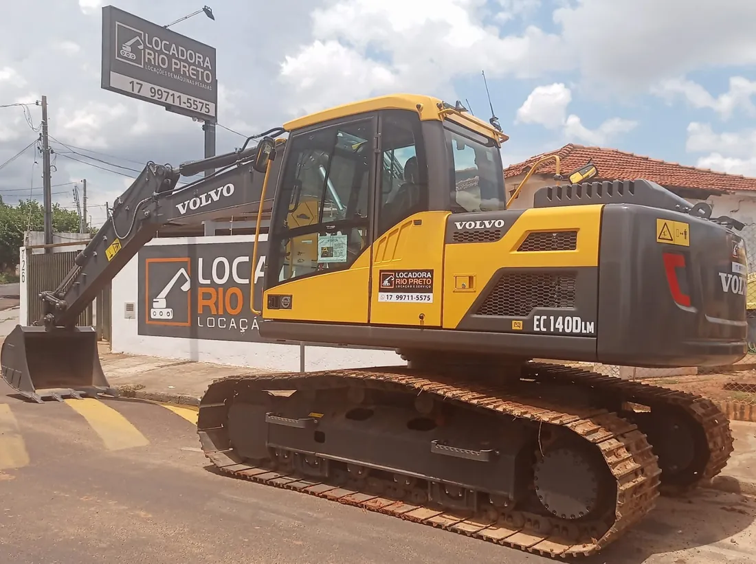Escavadeira hidráulica Volvo EC140DLM sobre esteiras, com adesivo da Locadora Rio Preto, diante da faixa da empresa