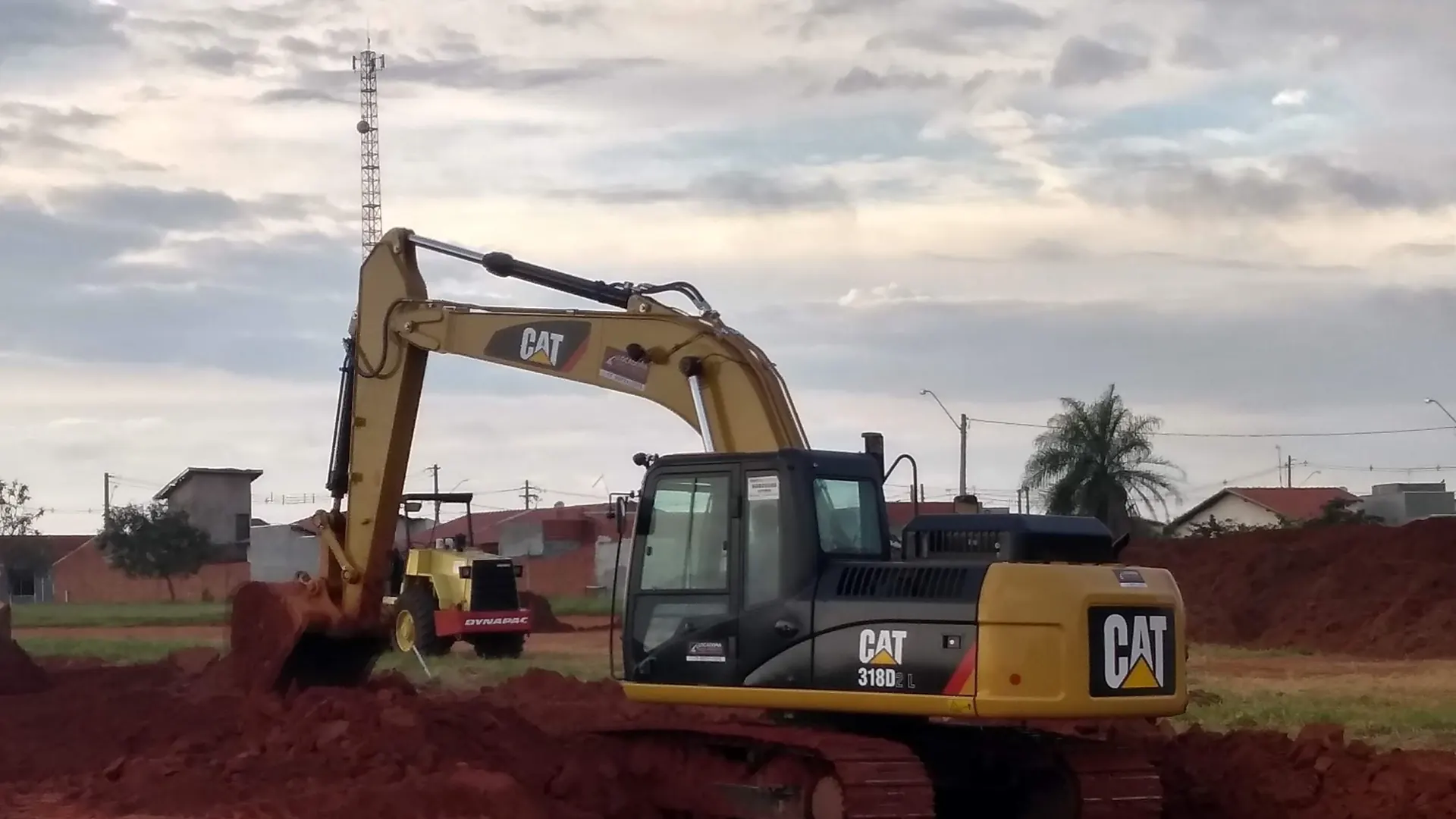 Escavadeira Caterpillar 318D2L abrindo vala em terra vermelha sob céu carregado, com um rolo compactador trabalhando ao fundo