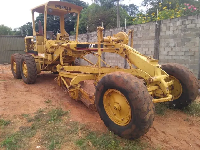 Motoniveladora Caterpillar 120 amarela, com a lâmina baixada, estacionada em pátio de terra vermelha