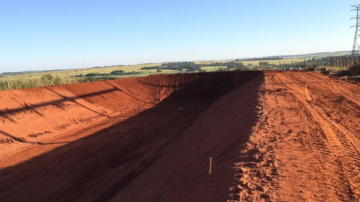 Bacia de contenção escavada ao lado de estrada de terra, com talude de terra vermelha