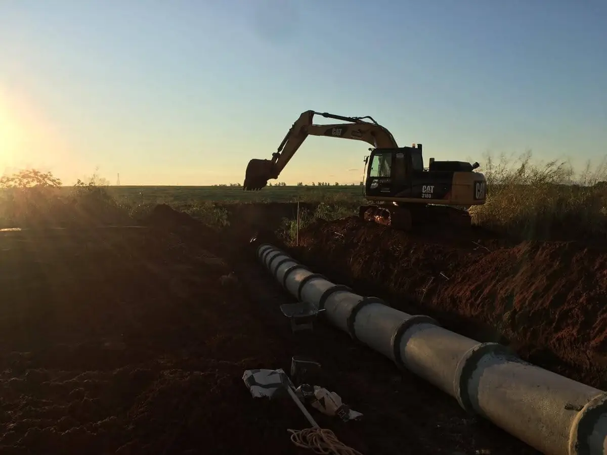 Escavadeira Caterpillar ao pôr do sol sobre vala com tubos de concreto assentados para drenagem