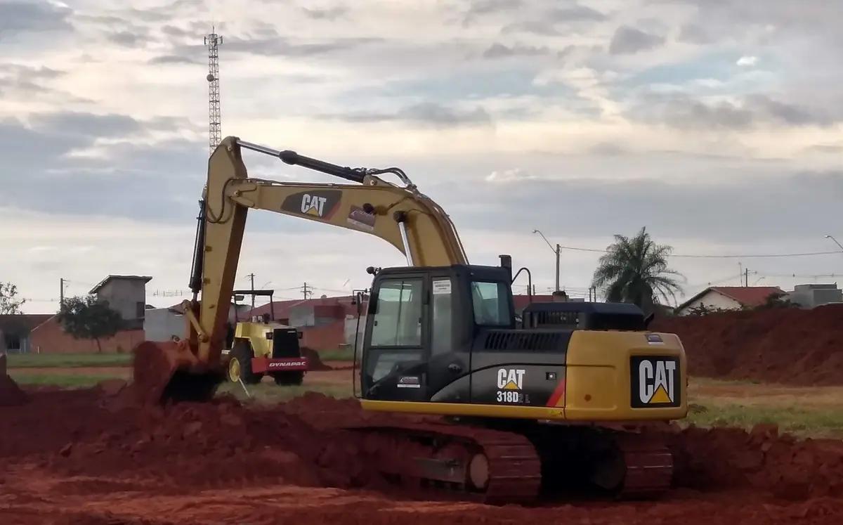 Escavadeira Caterpillar 318D2L abrindo vala em terra vermelha, sob céu nublado, com rolo compactador ao fundo