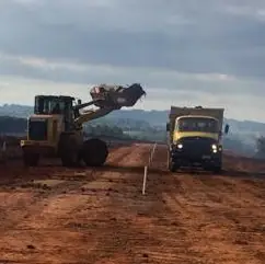 Pá carregadeira despejando terra na caçamba de um caminhão basculante em frente de obra