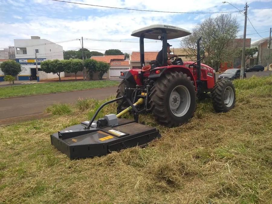 Trator Massey Ferguson vermelho com roçadeira acoplada trabalhando em terreno de grama alta, em área urbana