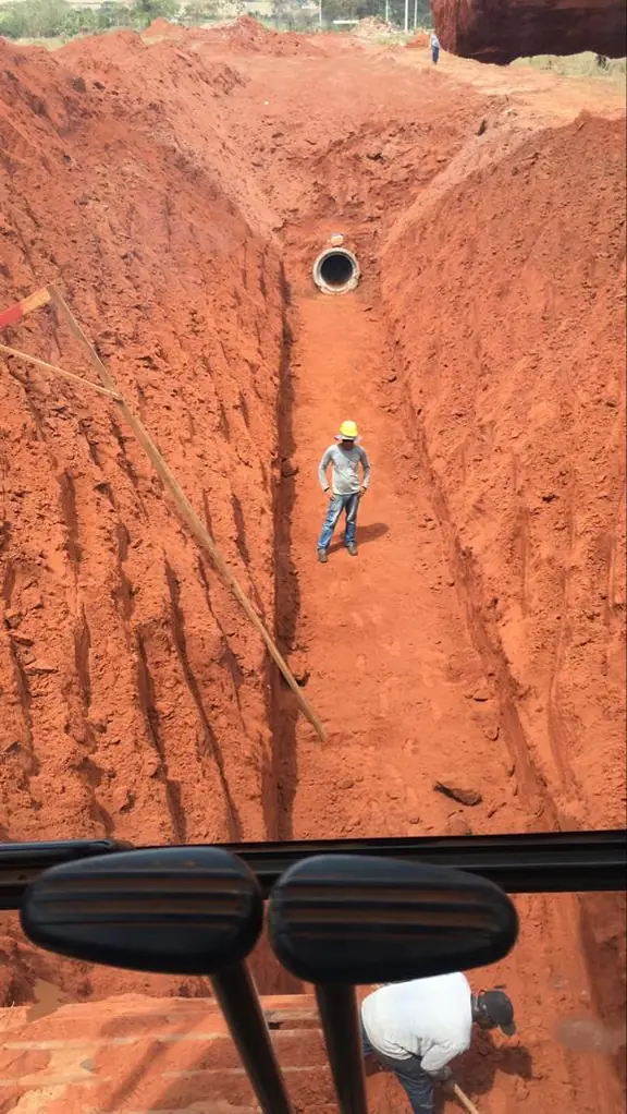 Vala profunda aberta em terra vermelha, com um trabalhador ao fundo dando a escala da profundidade