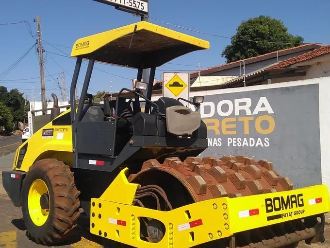 Rolo compactador pé de carneiro Bomag amarelo, de cabine aberta, diante do muro com a marca Locadora Rio Preto pintada