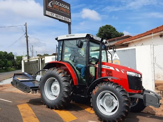 Trator Massey Ferguson 4707 de cabine fechada com roçadeira acoplada, diante da faixa da Locadora Rio Preto